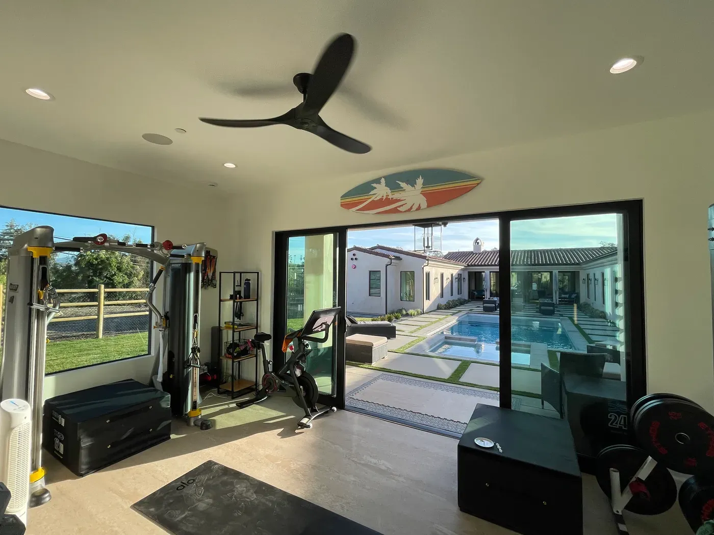 Gimnasio en casa con ventilador de techo negro de tres aspas y puertas de vidrio abiertas hacia una alberca