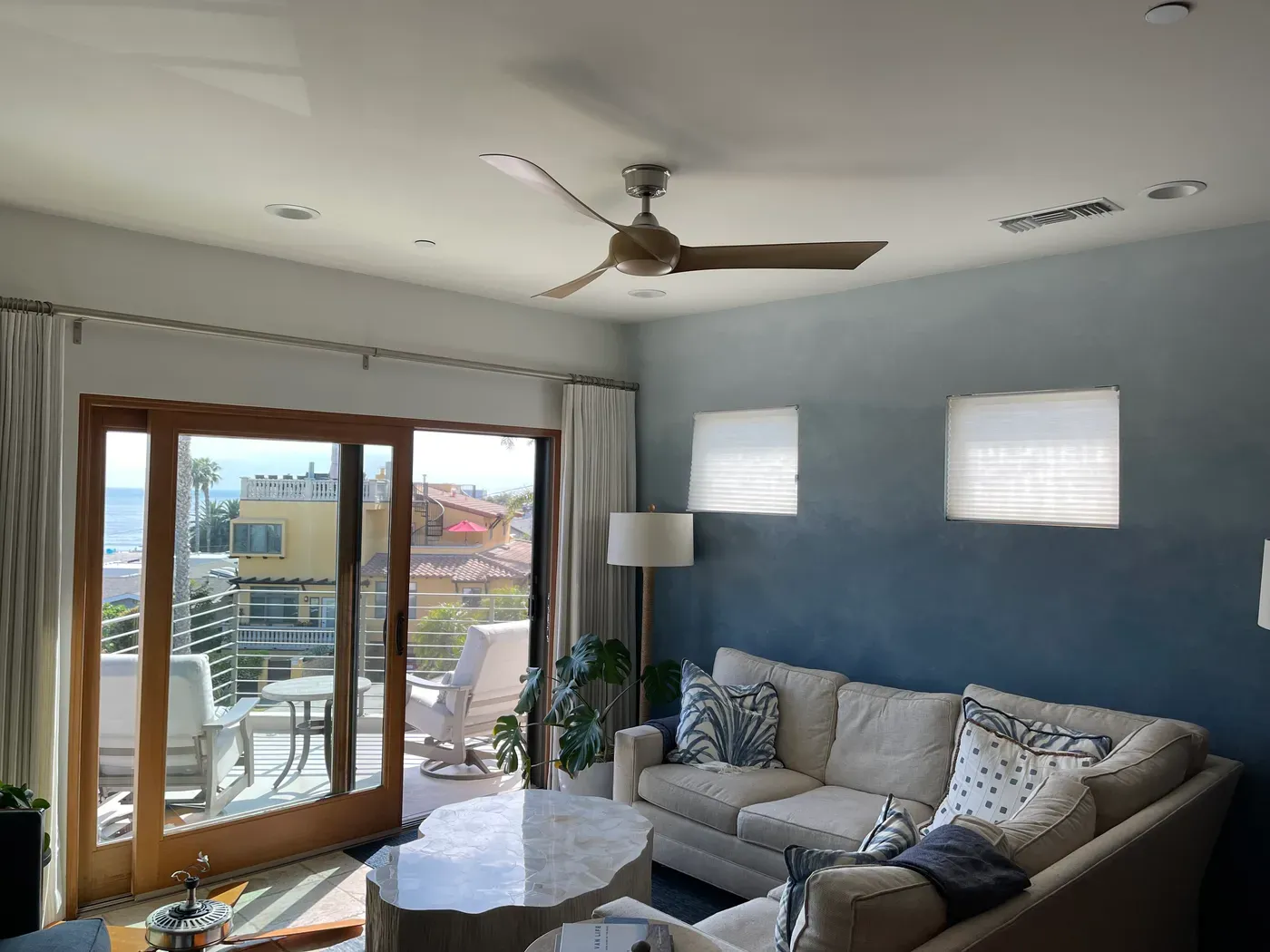 Sala junto a la costa con ventilador de techo moderno de tres aspas, sofá claro y puertas corredizas hacia una terraza con vista al mar